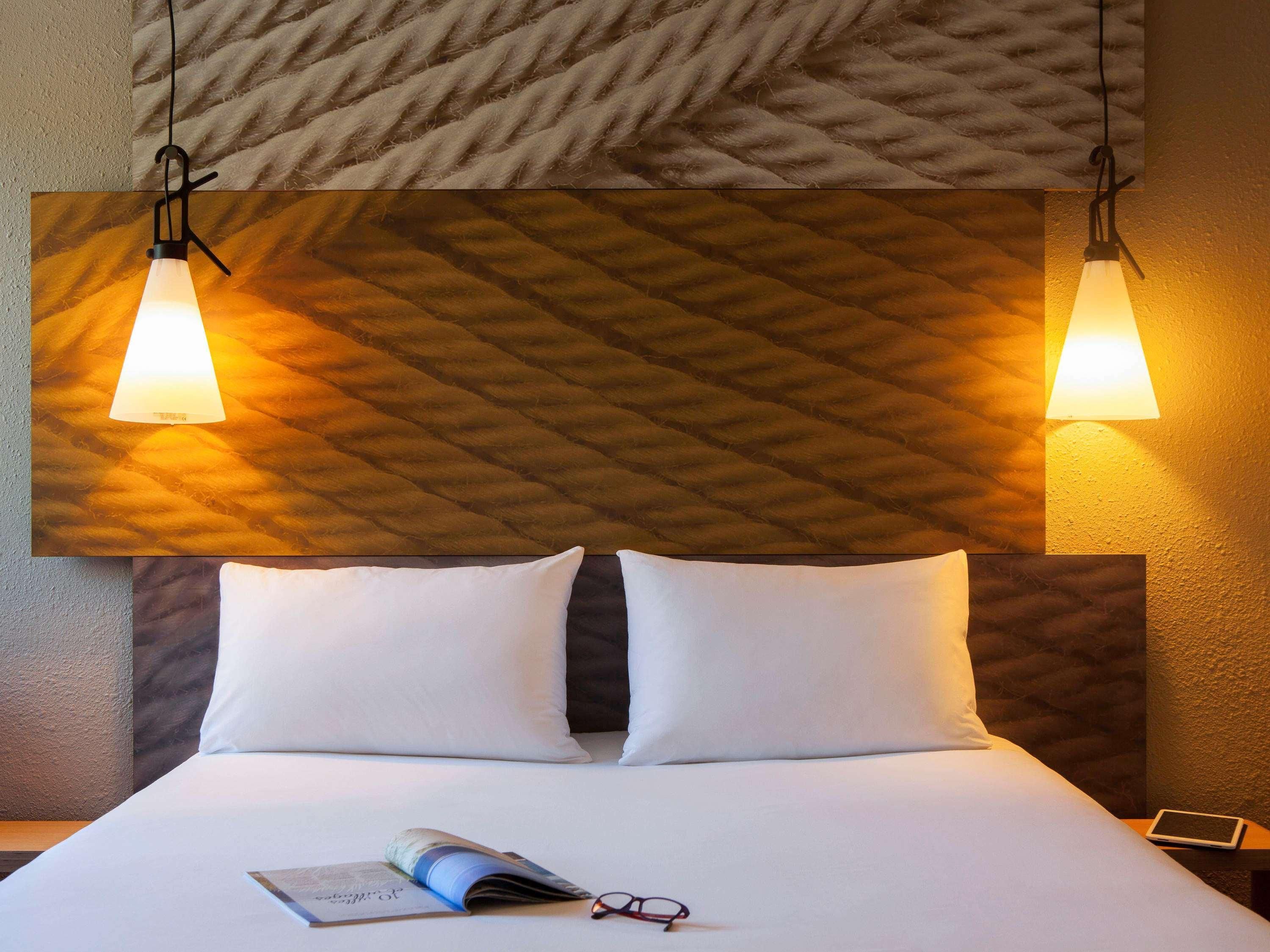 A hotel room with a bed, two bedside lamps, a headboard with a textured pattern, and a pillow.
