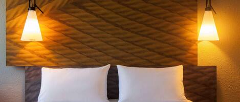 Una habitación de hotel con una cama, dos lámparas de noche, un cabecero con textura y una almohada.