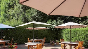 An outdoor patio area with round wooden tables and chairs, large umbrellas, and potted plants. - Ibis Jouy en Josas Velizy (Jouy-en-Josas)