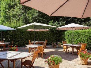 An outdoor patio area with round wooden tables and chairs, large umbrellas, and potted plants. - Ibis Jouy en Josas Velizy (Jouy-en-Josas)