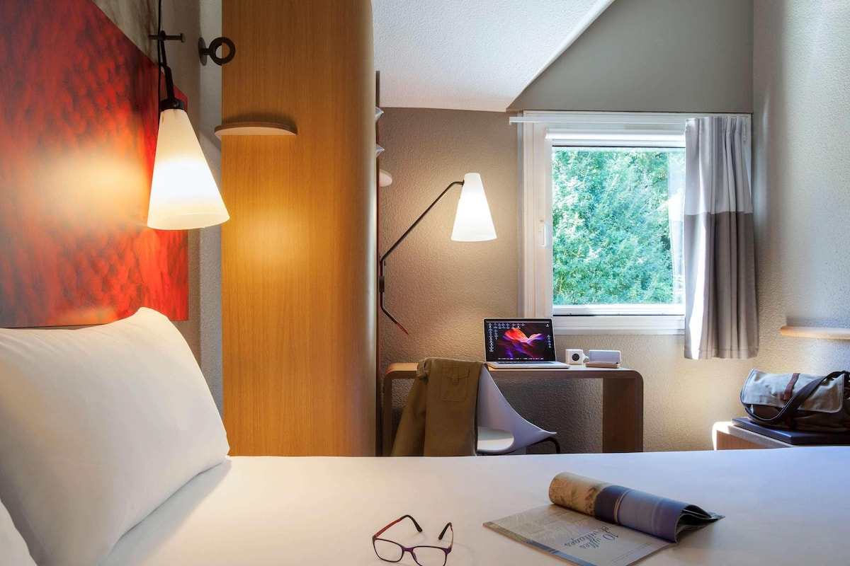Ein Hotelzimmer mit einem Bett, einem Schreibtisch mit Laptop, einem Fenster mit Vorhängen und einer Wandlampe.