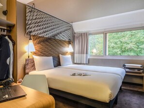 A hotel room with a bed, a desk with a laptop, a chair, a window with curtains, and a wall-mounted lamp. - Ibis Jouy en Josas Velizy (Jouy-en-Josas)