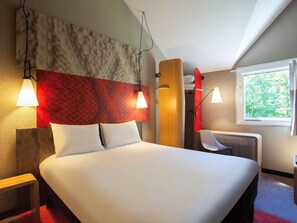A hotel room with a bed, two bedside lamps, a red accent wall, a wooden headboard, a desk with a chair, and a window with a view of trees. - Ibis Jouy en Josas Velizy (Jouy-en-Josas)