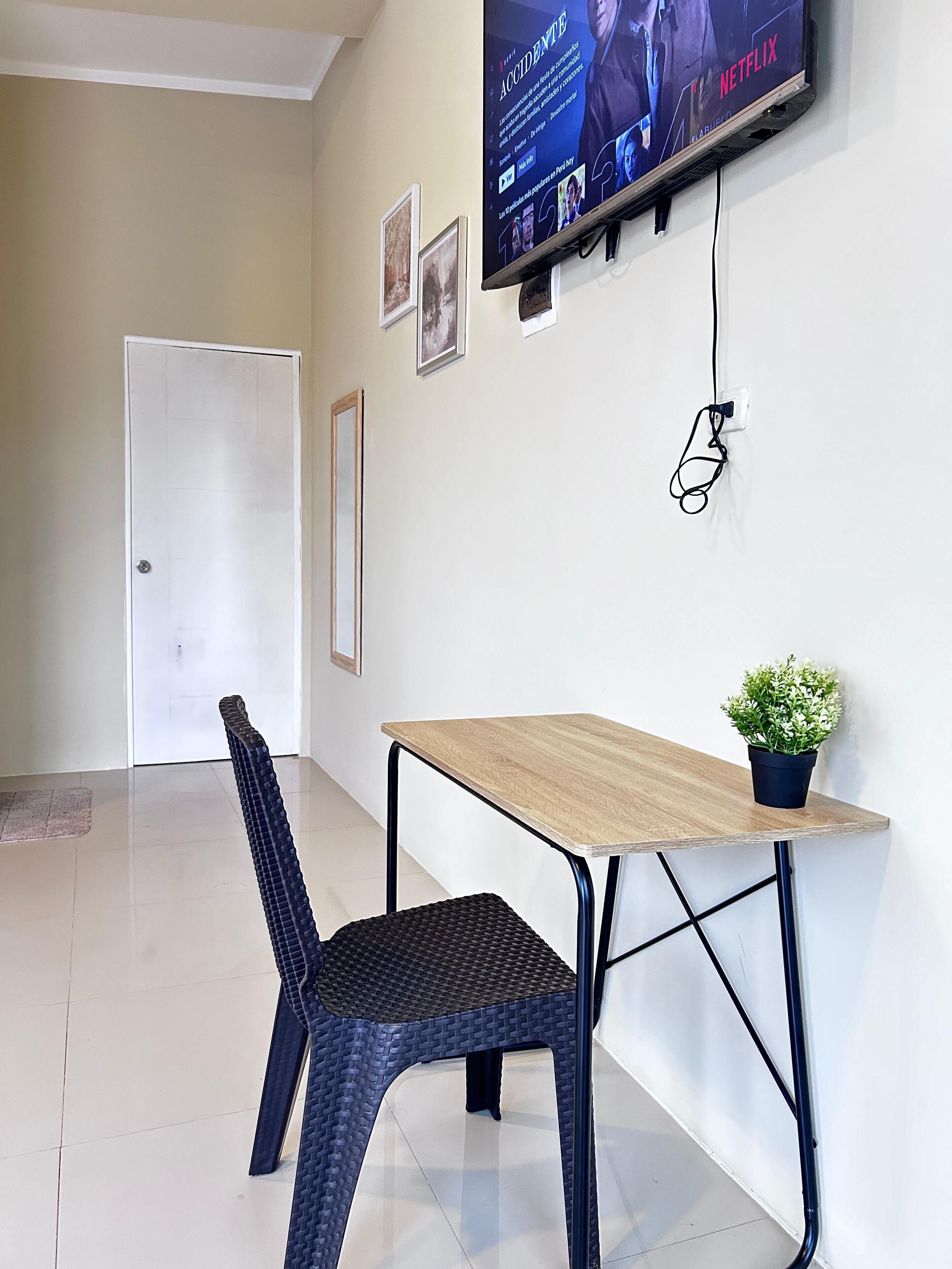 A room with a television mounted on the wall, a table with a potted plant, a chair, and a door.