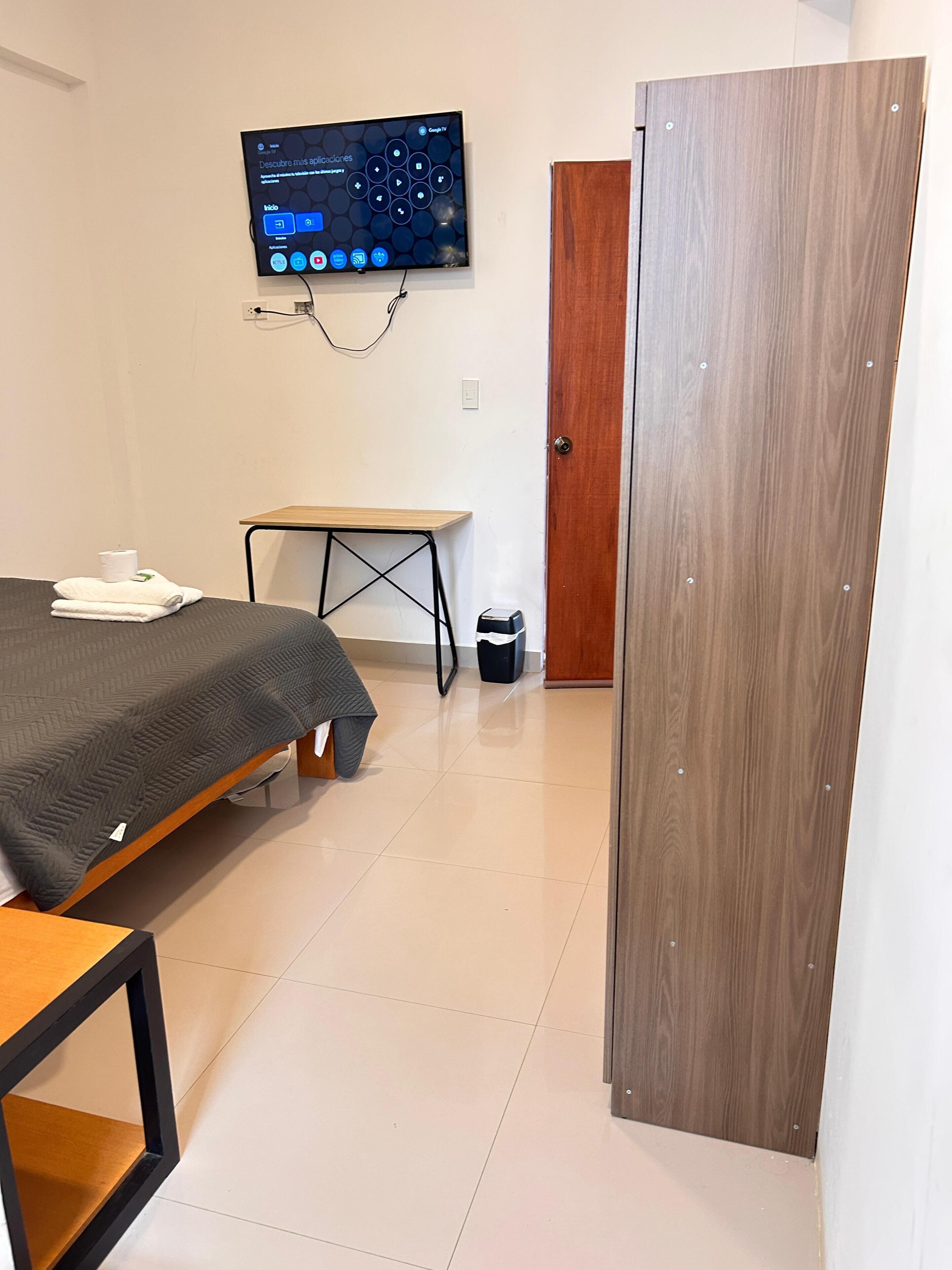 Una habitación con cama, escritorio, televisión instalada en la pared y puerta de madera.