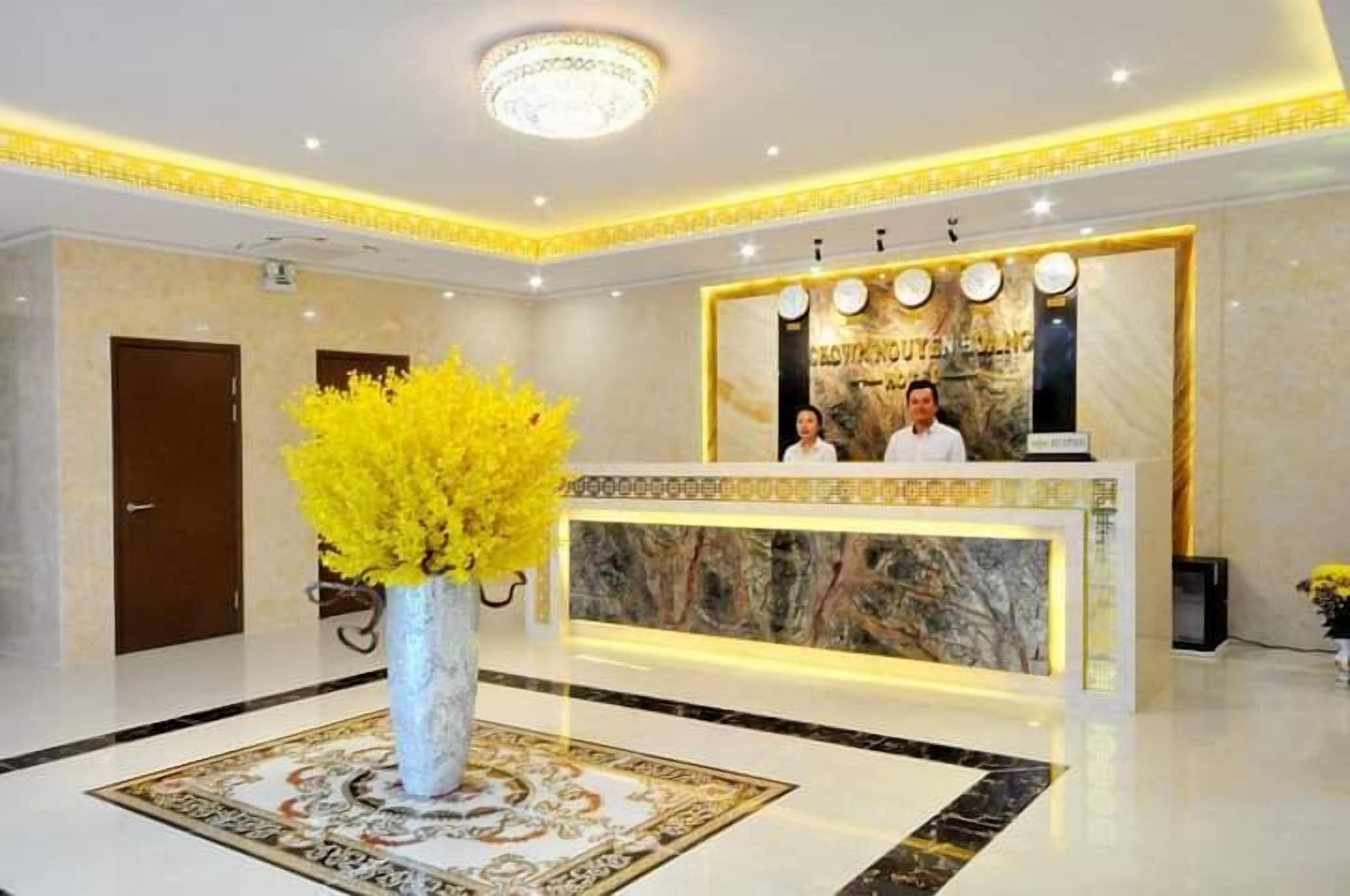 Zona de recepción de un hotel con un mostrador de mármol, dos recepcionistas, un jarrón grande con flores amarillas y un candelabro.