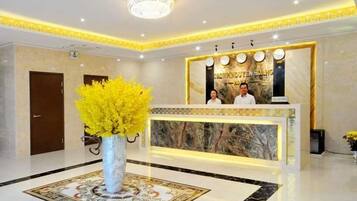Zona de recepción de un hotel con un mostrador de mármol, dos recepcionistas, un jarrón grande con flores amarillas y un candelabro.