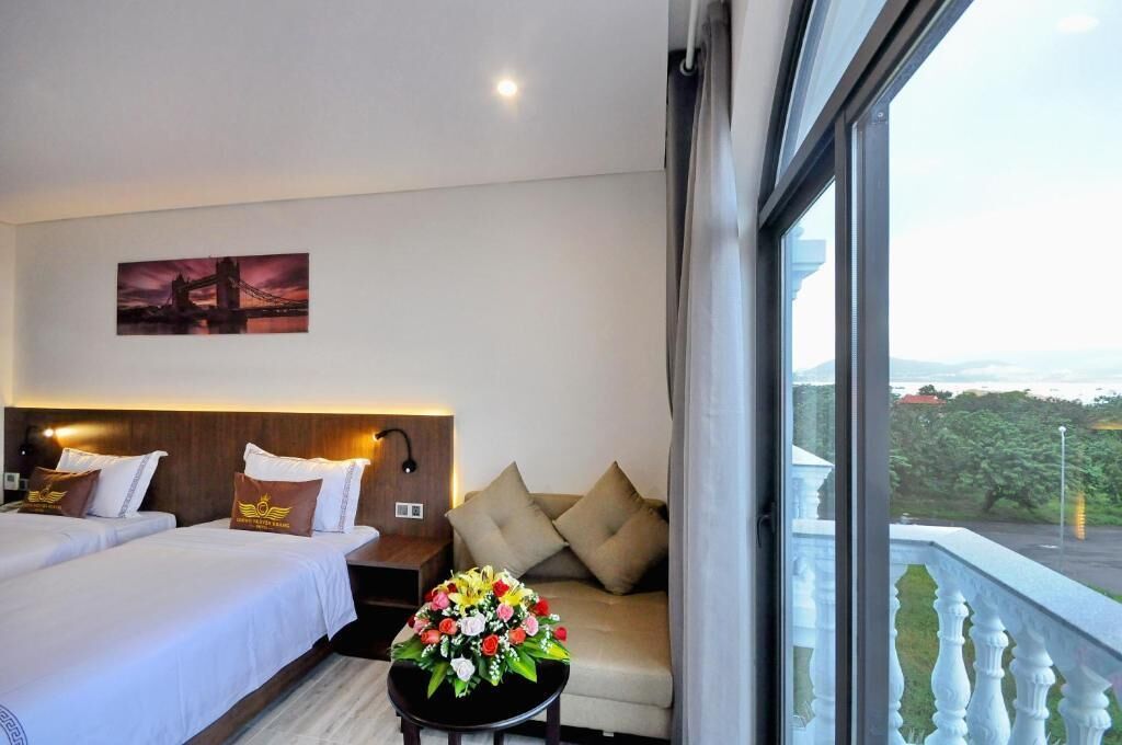 A hotel room with two beds, a sofa, a table with flowers, a painting on the wall, and a balcony with a view.