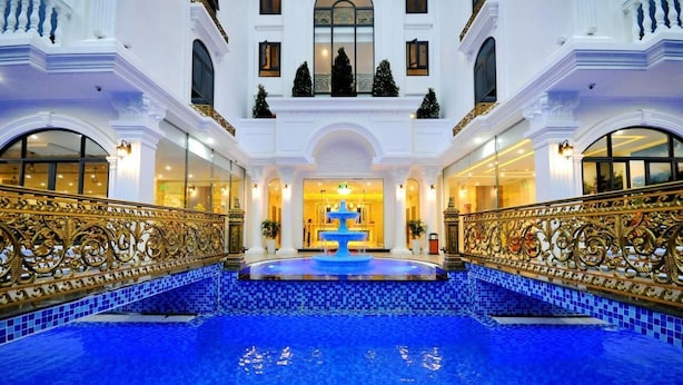 A grand hotel lobby with a central fountain, ornate gold railings, and a tiled blue pool.