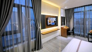 A modern hotel room with a large bed, a desk with a chair, a TV mounted on the wall, and large windows with curtains.