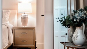 Ein Schlafzimmer mit Bett, Nachttisch mit Lampe, einem Spiegel und einer Vase mit Blumen.