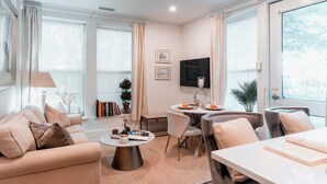 Ein modernes Wohnzimmer mit Sofa, Couchtisch und Essbereich. Große Fenster mit Vorhängen, ein Flachbildfernseher und ein Bücherregal sind ebenfalls vorhanden.