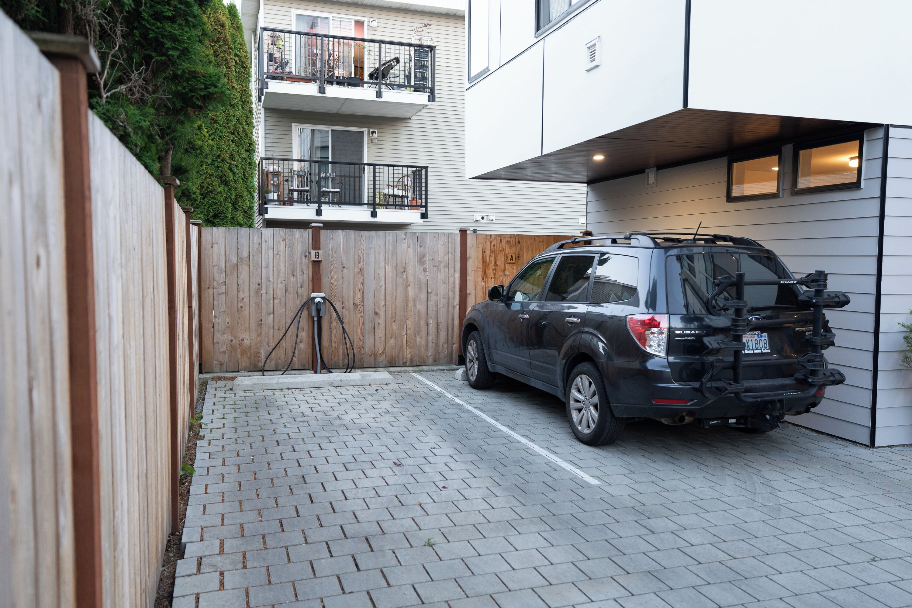 Un’area di parcheggio con superficie pavimentata, una recinzione in legno e un SUV nero parcheggiato vicino a un edificio con un balcone.