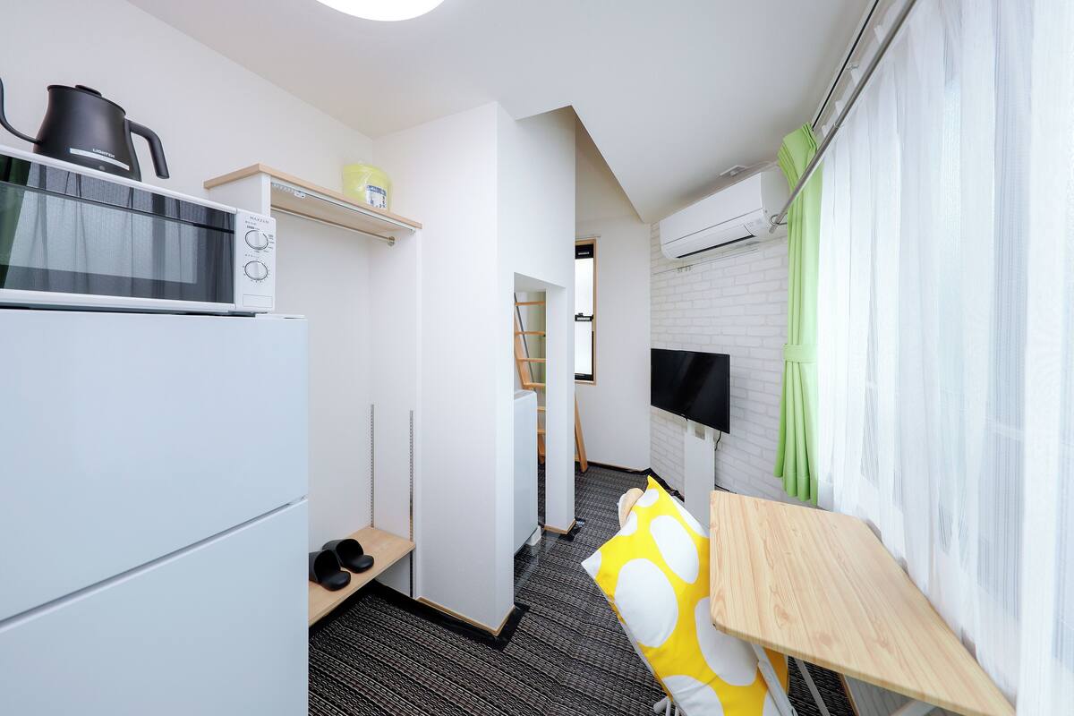 A compact kitchen with a microwave, kettle, and shelf with a yellow container. A small table with a yellow and white polka dot cushion is visible.