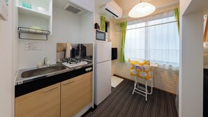 A compact kitchen with a sink, stove, microwave, and refrigerator. There is a small stool and a window with curtains. - Zenith Shinjuku Hinoki Building A (Tokyo)