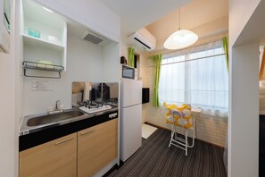 A compact kitchen with a sink, stove, microwave, and refrigerator. There is a small stool and a window with curtains. - Zenith Shinjuku Hinoki Building A (Tokyo)