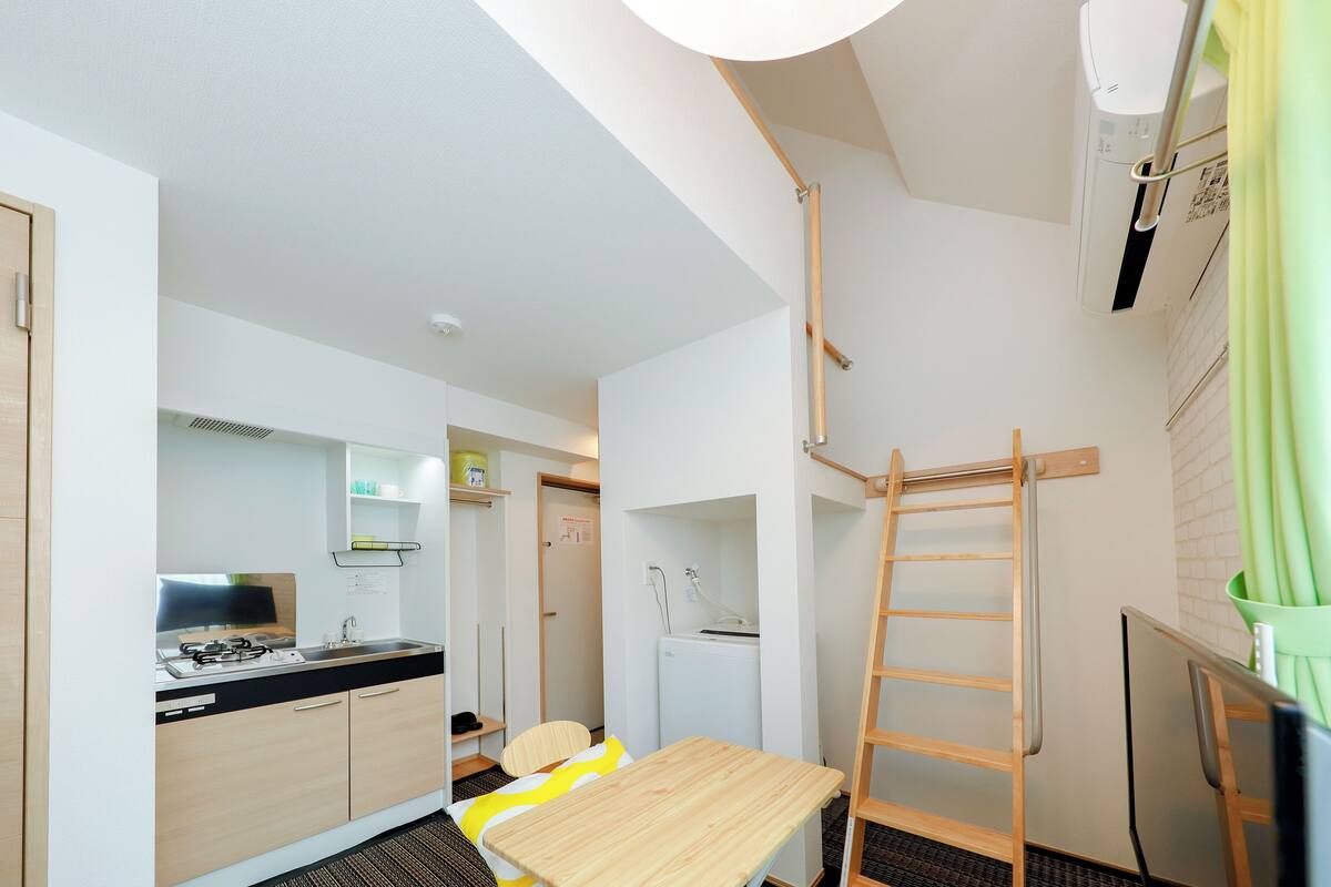 A compact kitchen with a stove, sink, and a small refrigerator. A wooden ladder is leaning against the wall, and a chair is placed at a table.