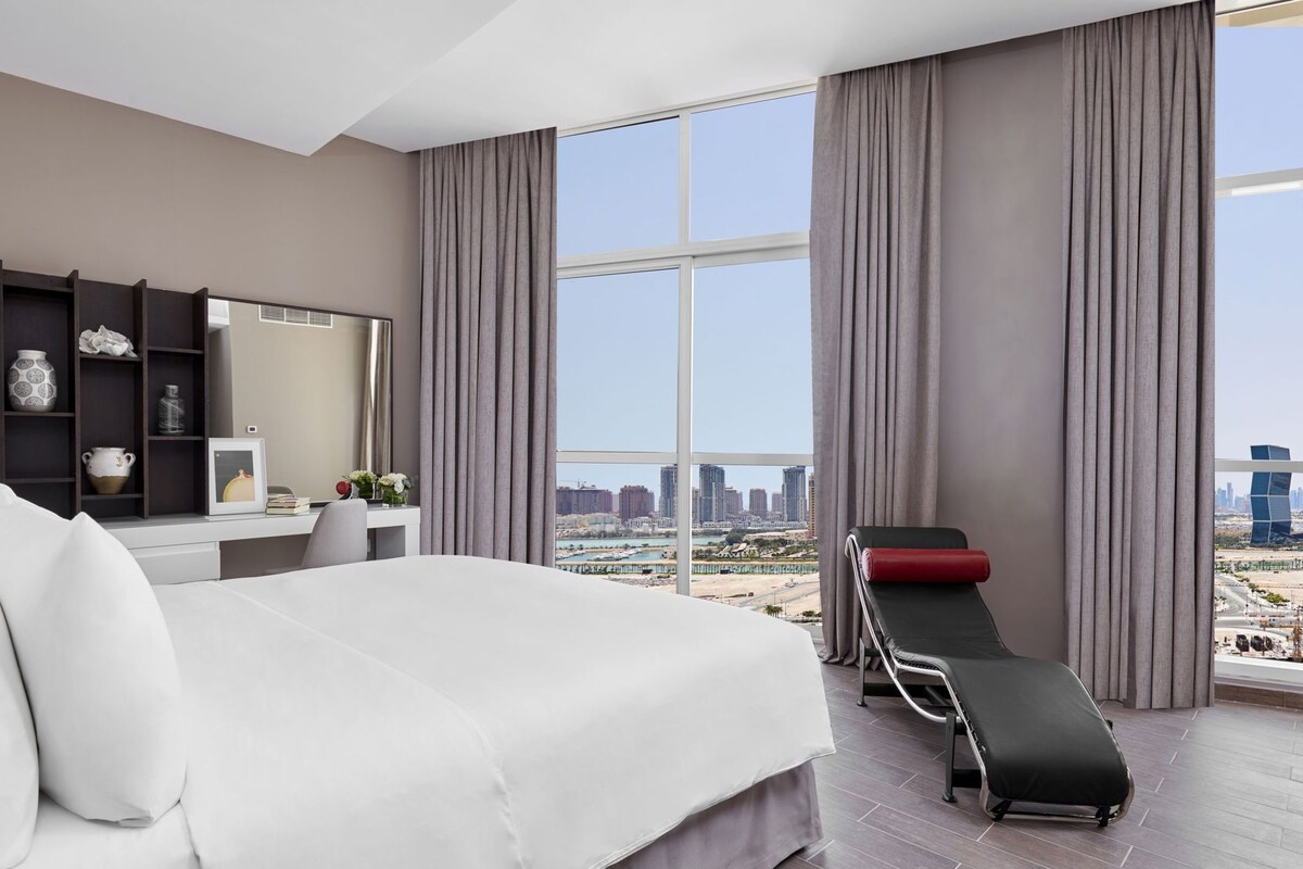 A modern hotel room with a large bed, a desk with a mirror, a bench with a red cushion, and a city view through large windows.