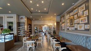 Un café espacioso con sillas y mesas blancas, una vitrina con pasteles y una pared decorada con cuadros.