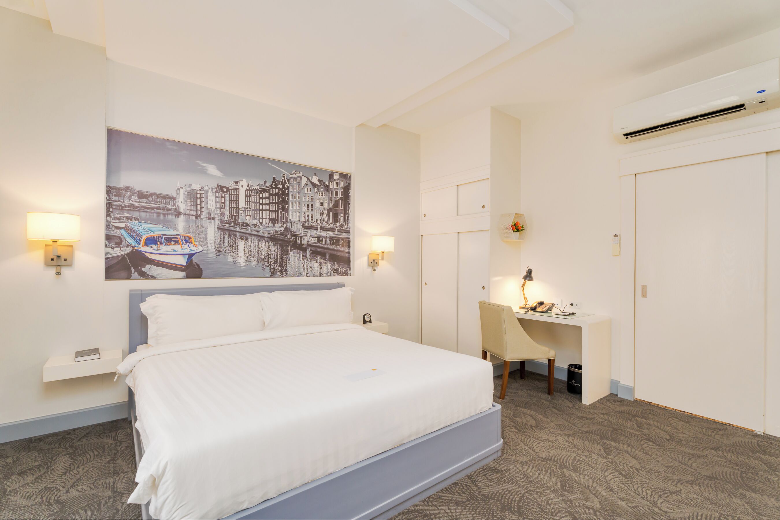 A hotel room with a large bed, a desk, two wall-mounted lamps, and a cityscape painting on the wall.
