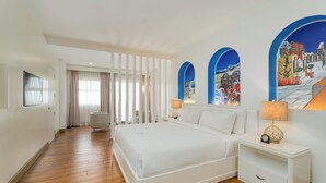Habitación de hotel con una cama grande, mesitas de noche blancas, piso de madera y vistas a un pueblo costero a través de ventanales arqueados.