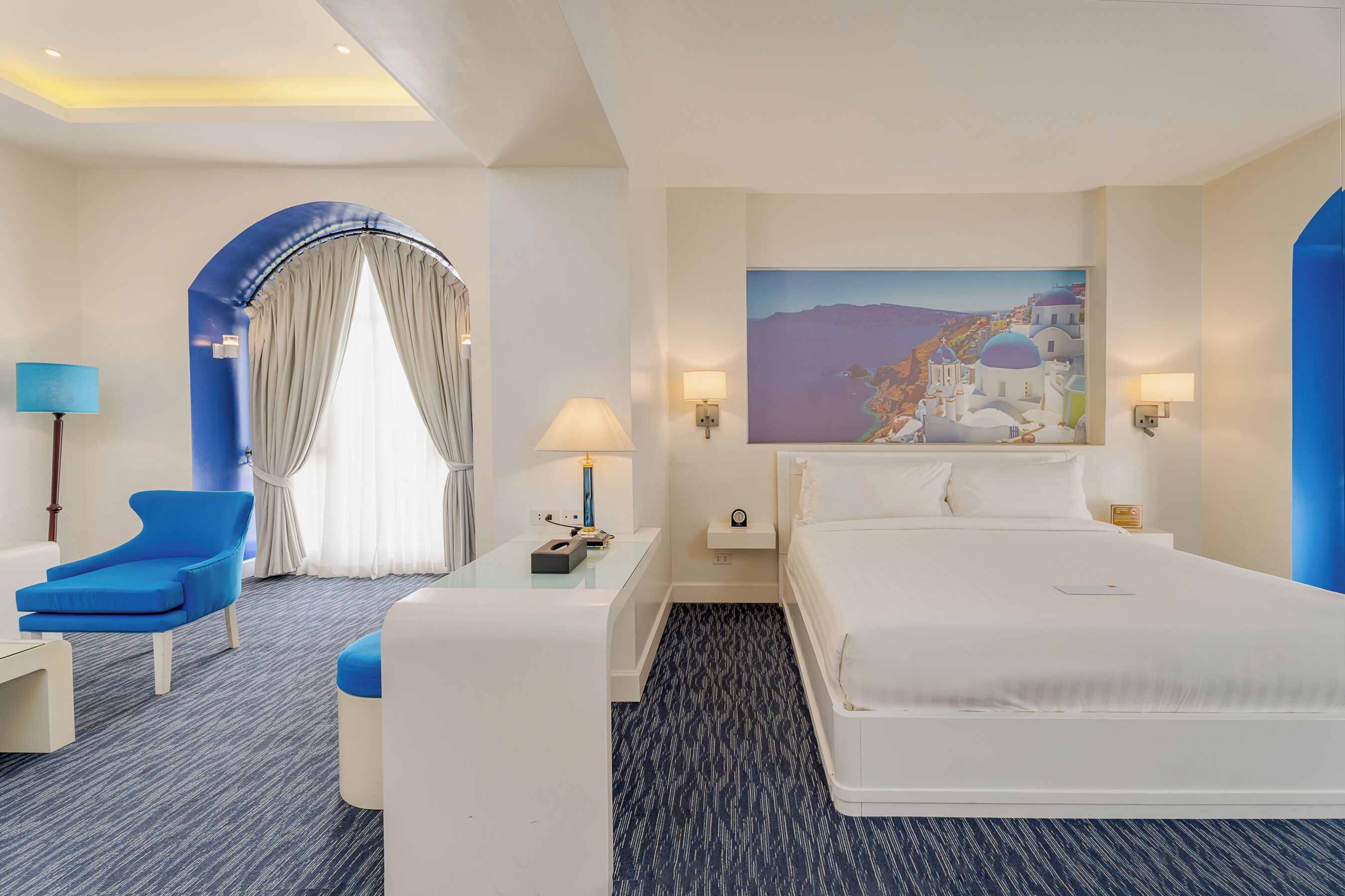 A hotel room with a large bed, a desk with a chair, a painting of a coastal scene, and a window with curtains.