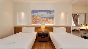A hotel room with two beds, a nightstand with a lamp, a painting of a coastal town, and a bathroom with a mirror and towel rack.