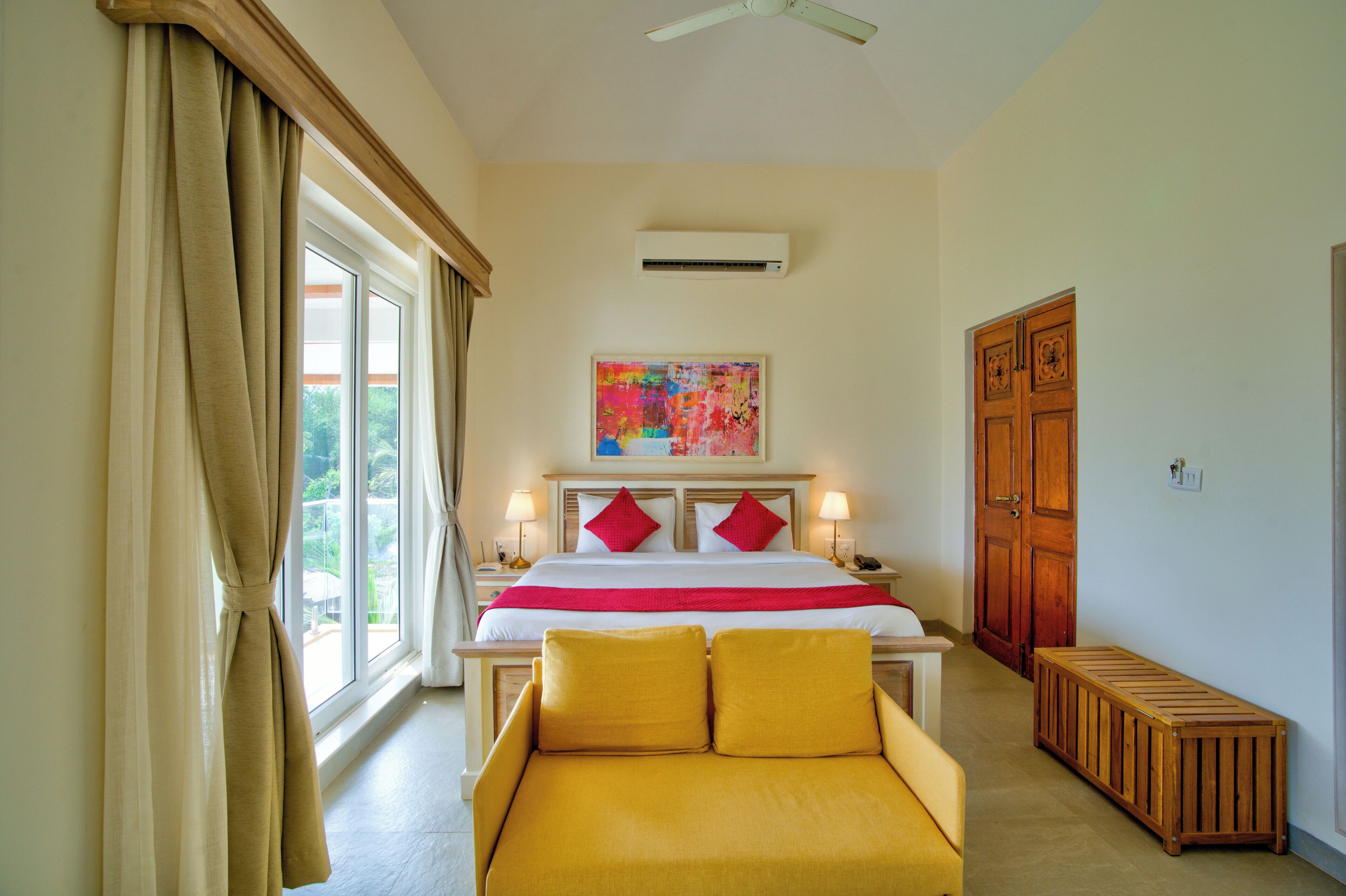 A hotel room with a bed, bedside lamps, a bench, a window with curtains, an air conditioning unit, and a door.