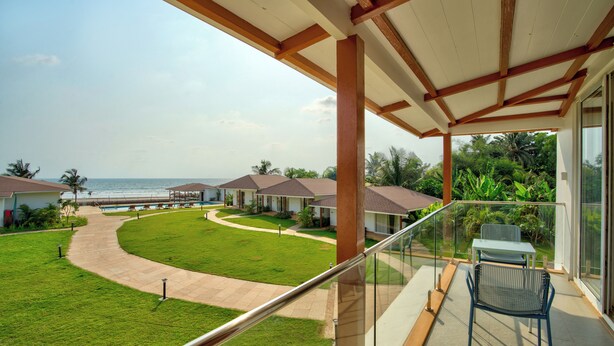A balcony with a view of the ocean, a grassy lawn, and a pathway leading to a building with a pool. - The Goa Beach Resort Morjim (Morjim)