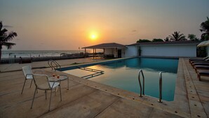 A swimming pool with lounge chairs and a table, overlooking the ocean at sunset. - The Goa Beach Resort Morjim (Morjim)