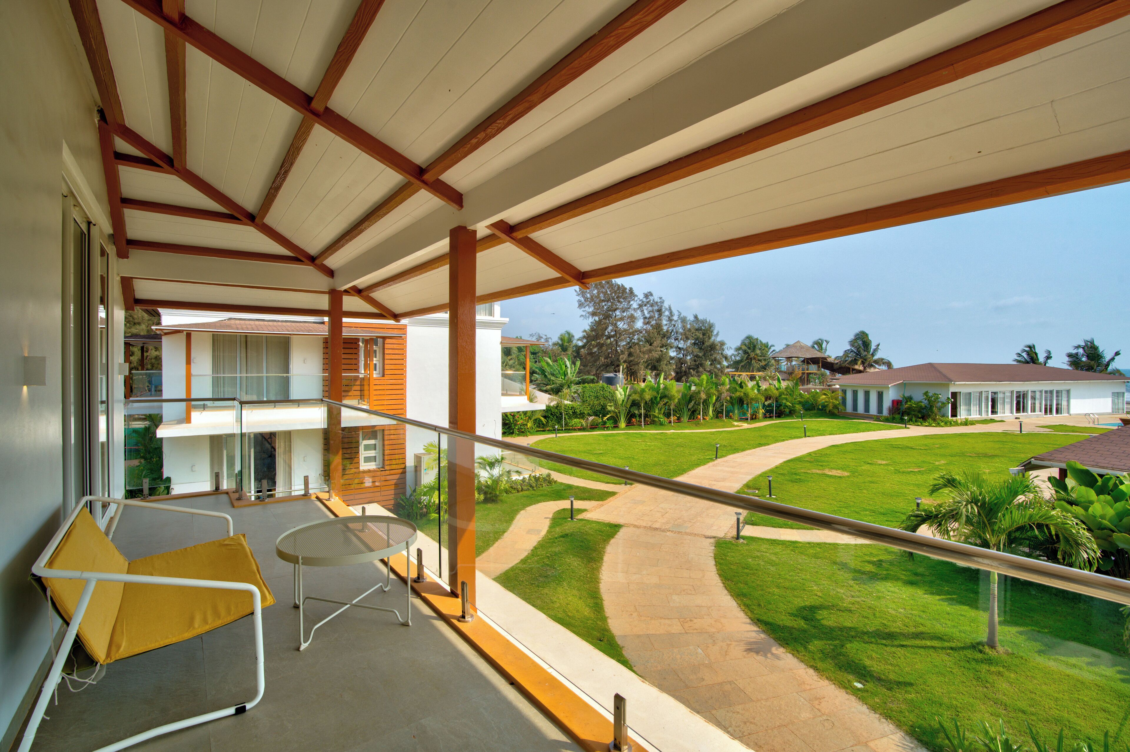 A balcony with a yellow chair and a small table, overlooking a well-maintained lawn and a building with a red roof.