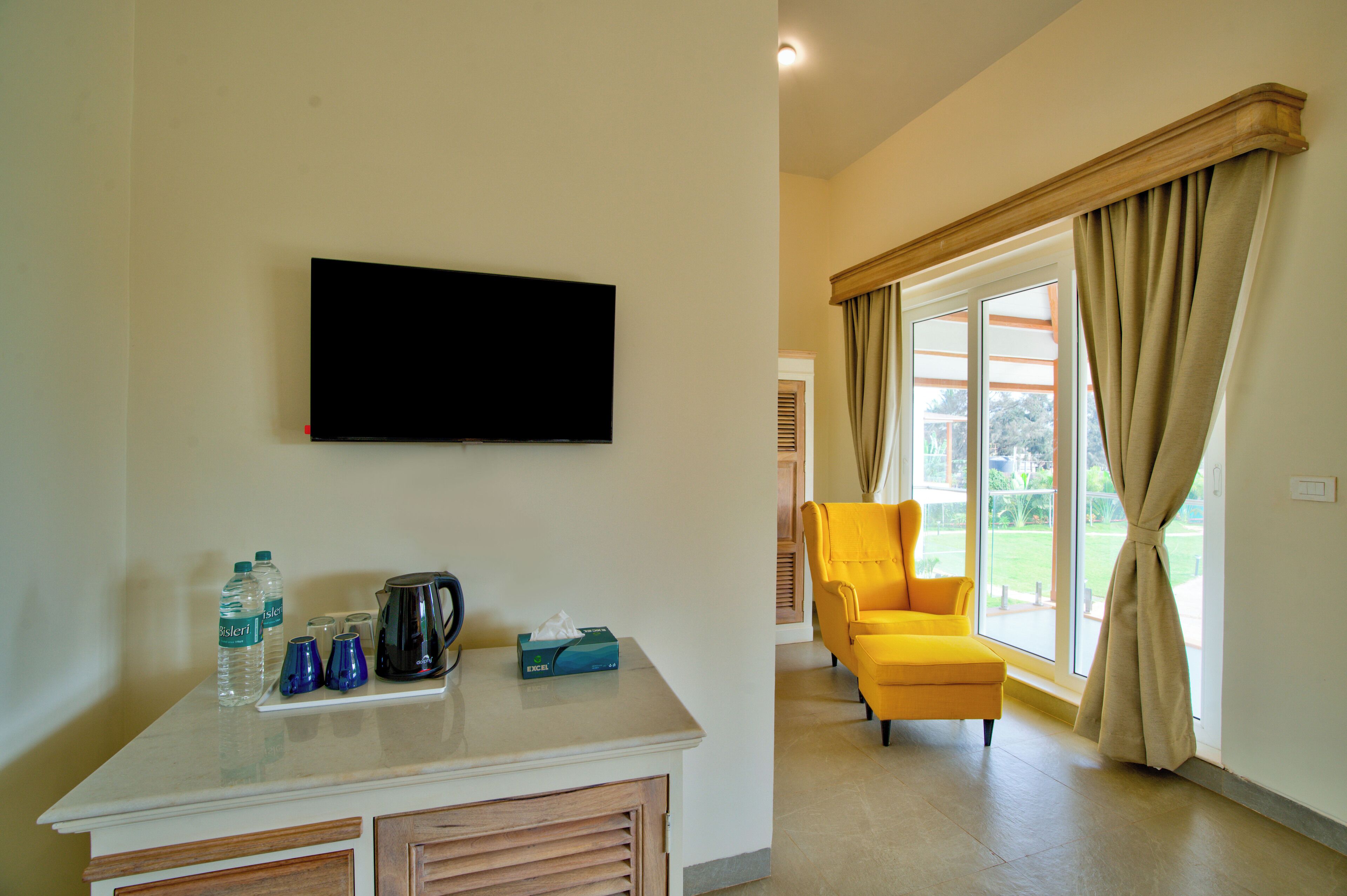 Habitación con televisión de pantalla plana montada en la pared, una mesita con cafetera, botellas de agua y una caja de pañuelos. Un sillón amarillo se encuentra cerca de un ventanal con cortinas color beige.