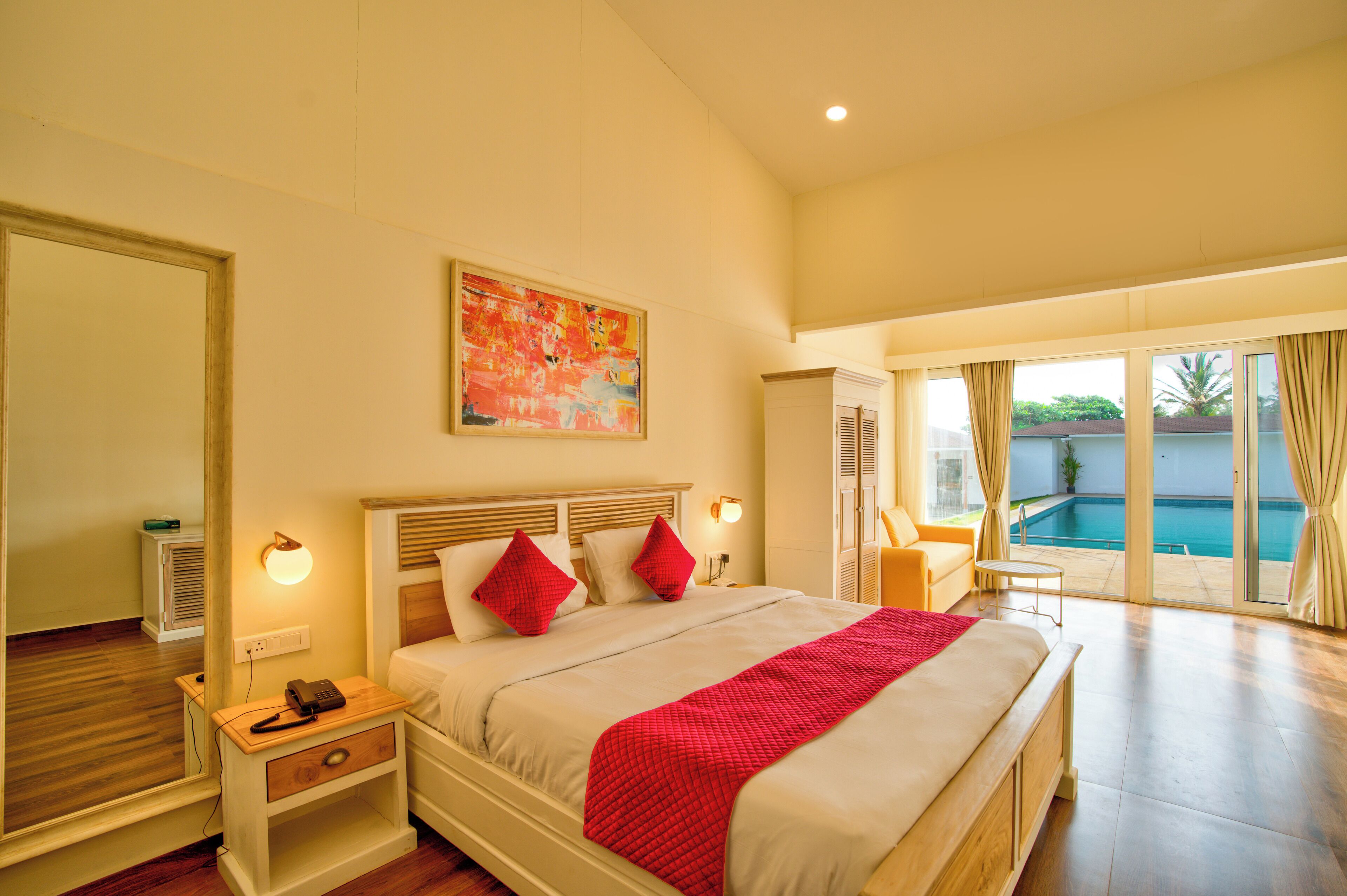 A spacious bedroom with a large bed, a bedside table with a lamp, a painting on the wall, and a view of a swimming pool through the glass doors.