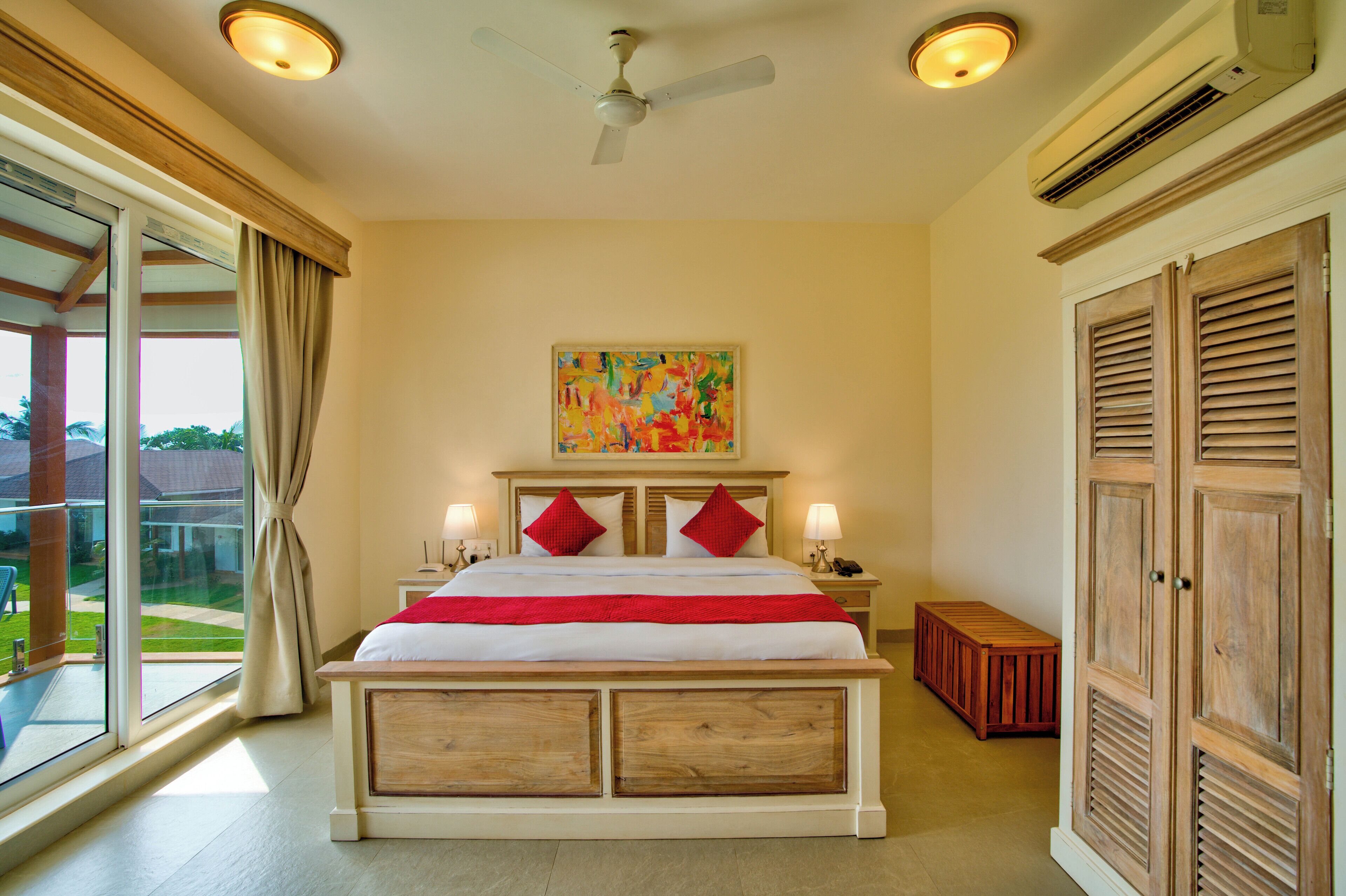 A bedroom with a large bed, a ceiling fan, an air conditioning unit, a nightstand with a lamp, a wooden bench, and a view of the outdoors.