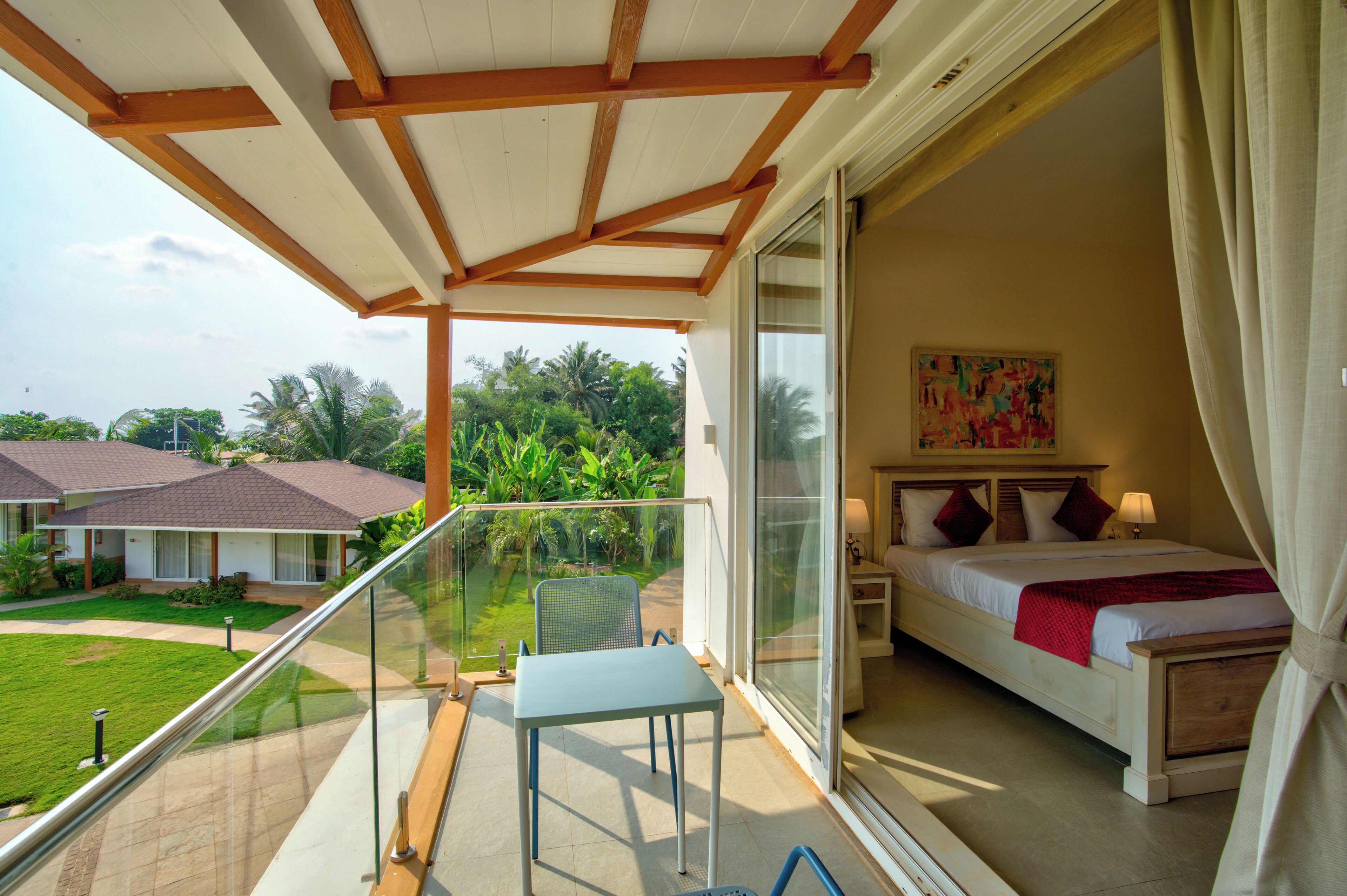 A balcony with a table and chairs, a bed with a red blanket, and a painting on the wall.