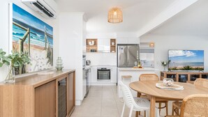 Et moderne køkken med hvide skabe, et rustfrit stål køleskab og et spiseområde med et træbord og fletstole.