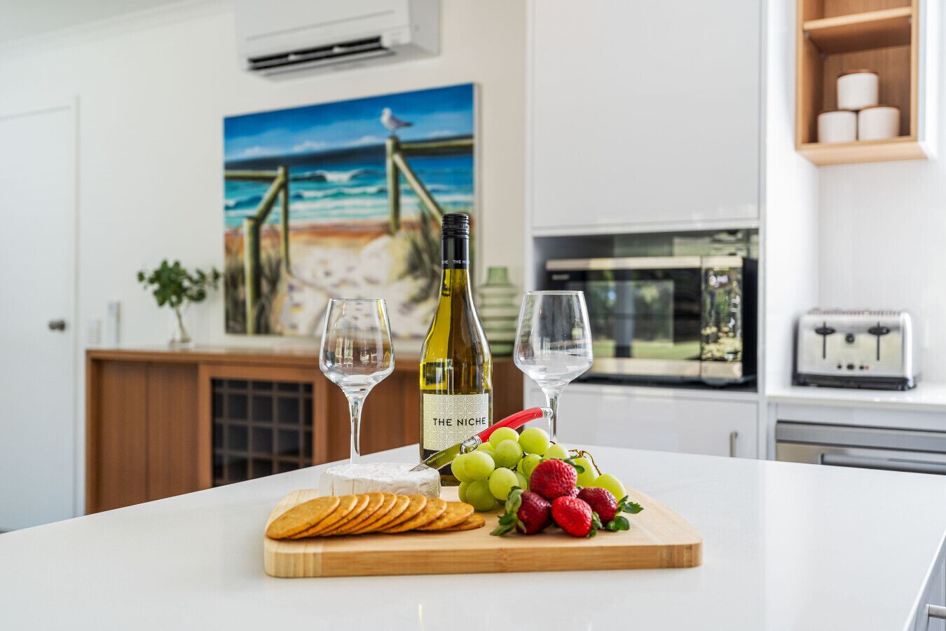 A kitchen counter with a bottle of wine, two glasses, a wooden cutting board with grapes and strawberries, and a painting of a beach scene in the background.