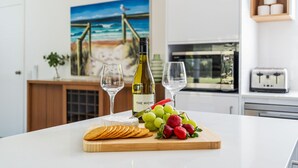 En køkkenbordplade med en vinflaske, to glas, en træskærebræt med druer og jordbær, og et maleri af en strandscene i baggrunden.