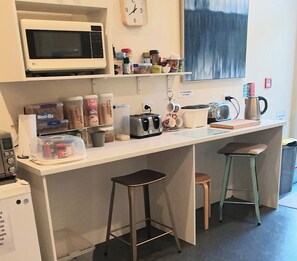 Eine kompakte Küchenzeile mit Mikrowelle, Toaster und Kaffeemaschine. Es gibt Aufbewahrungsbehälter und eine Uhr an der Wand. Zwei Hocker stehen an der Arbeitsplatte.