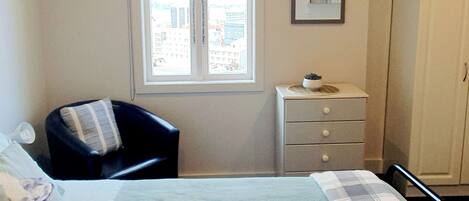 A bedroom with a bed, a chair, a dresser, and a window with a view of buildings.