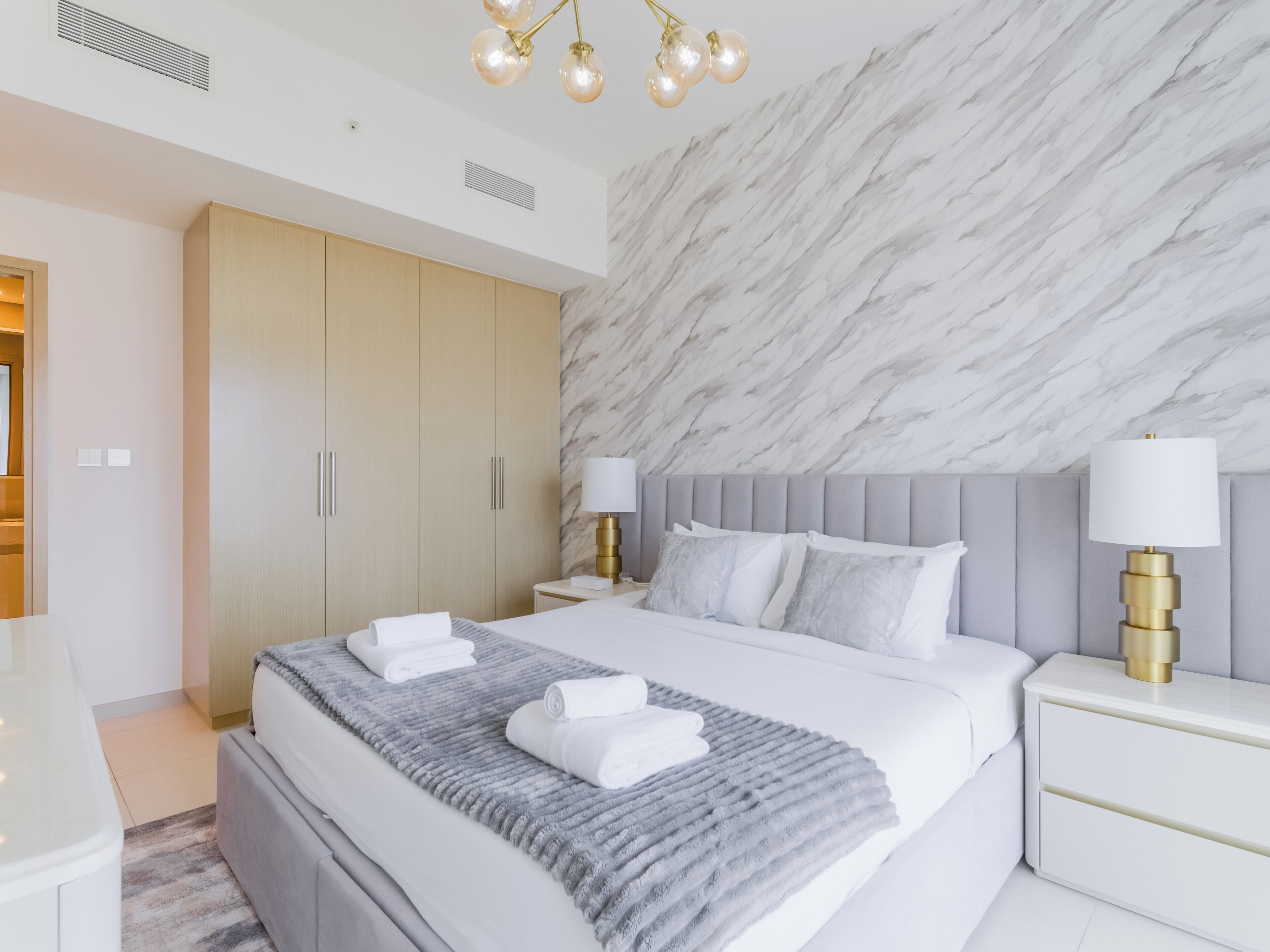 Een moderne hotelkamer met een wand in marmerlook, een groot bed met witte lakens, twee nachtkastjes met lampen en een kledingkast.