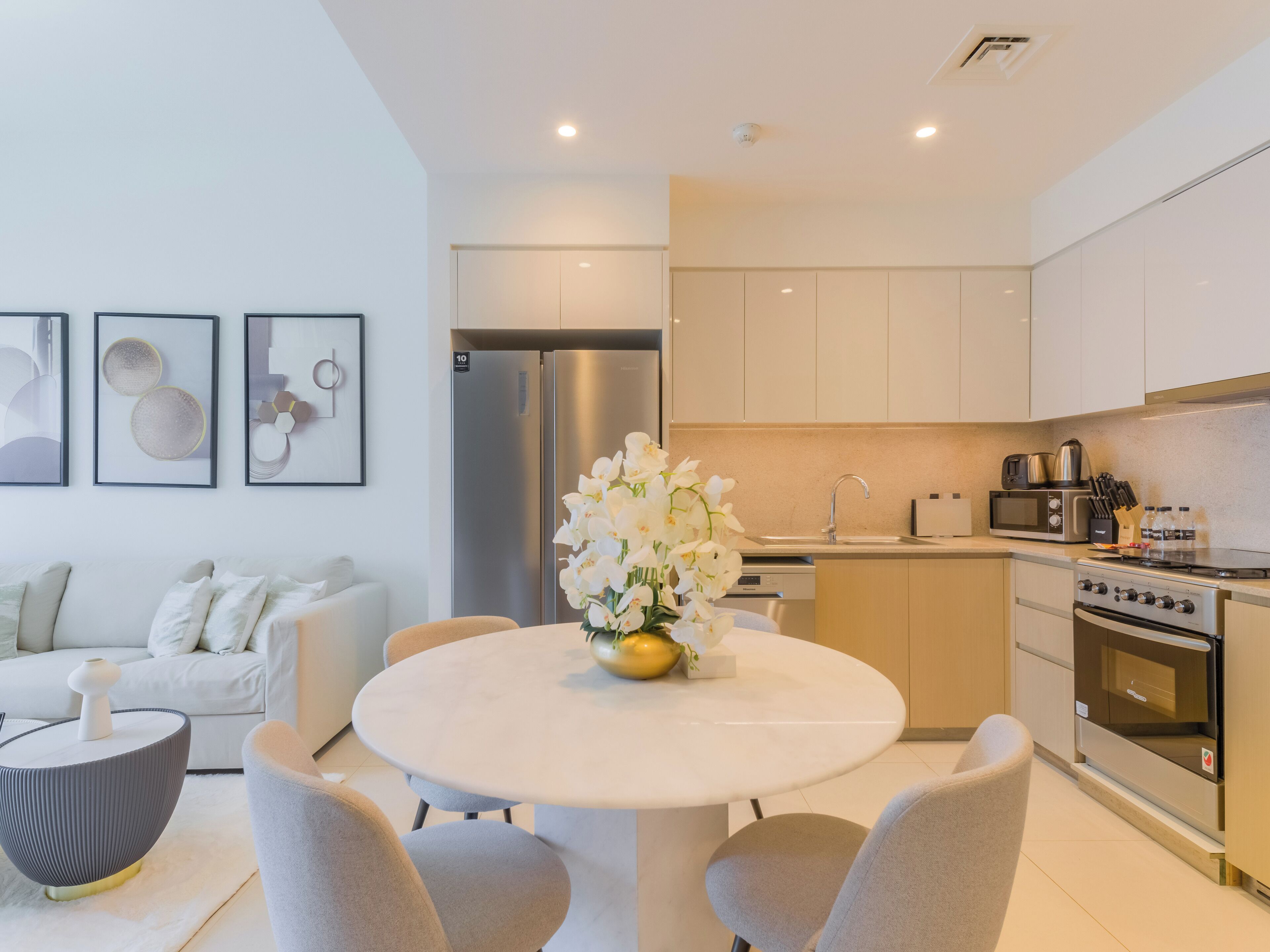 Una cocina moderna con electrodomésticos de acero inoxidable, un comedor con una mesa redonda y sillas, y una sala de estar con un sofá y objetos decorativos.