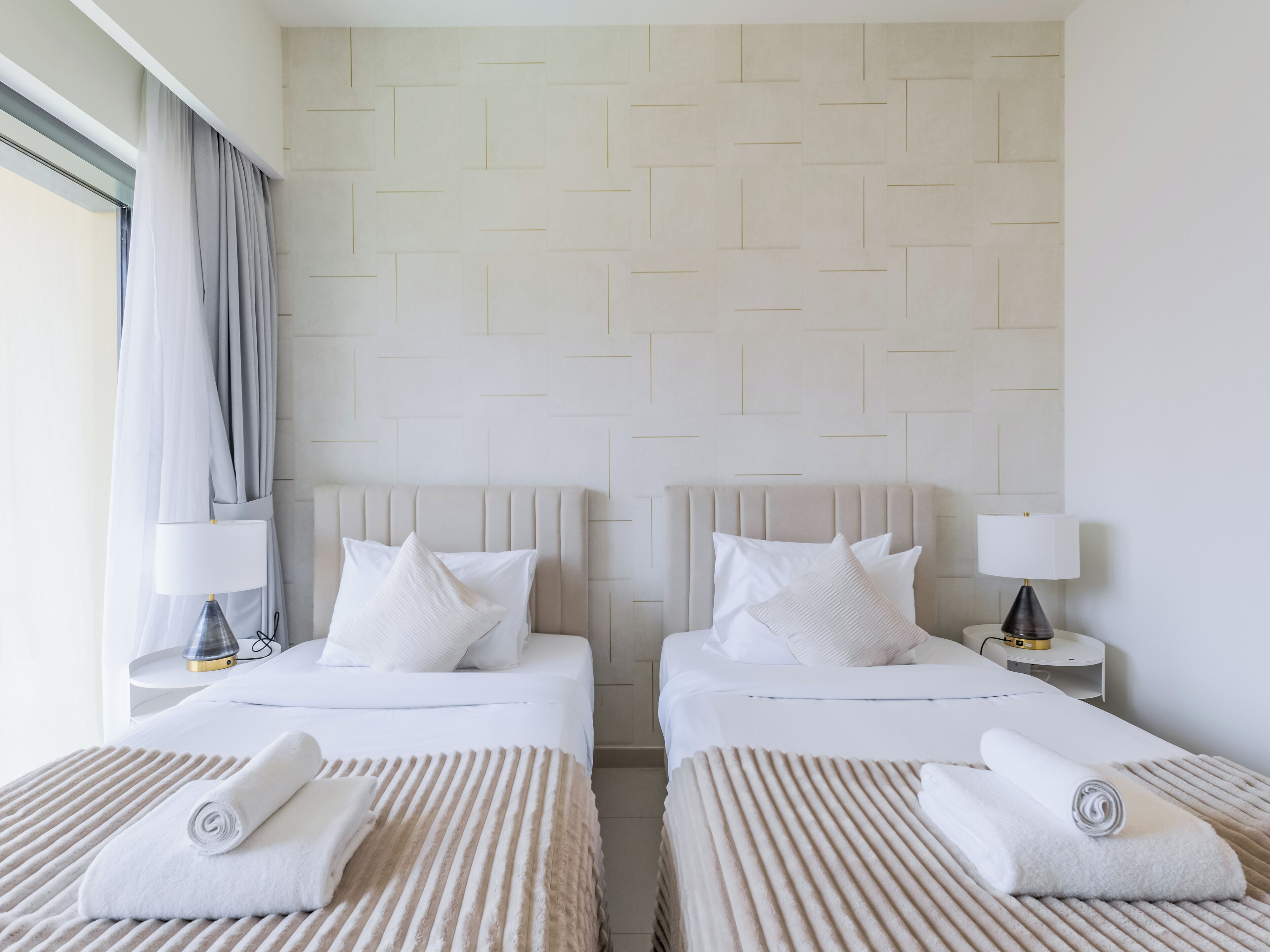Habitación de hotel con dos camas, un ventanal con cortinas, una mesita de noche con lámpara y una pared con diseño geométrico.