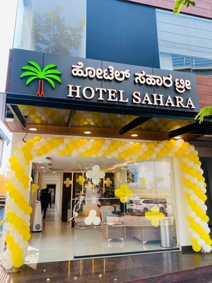 A hotel entrance with a sign in multiple languages, a palm tree logo, and a yellow and white balloon arch. - Hotel Sahara Tree (Bengaluru)