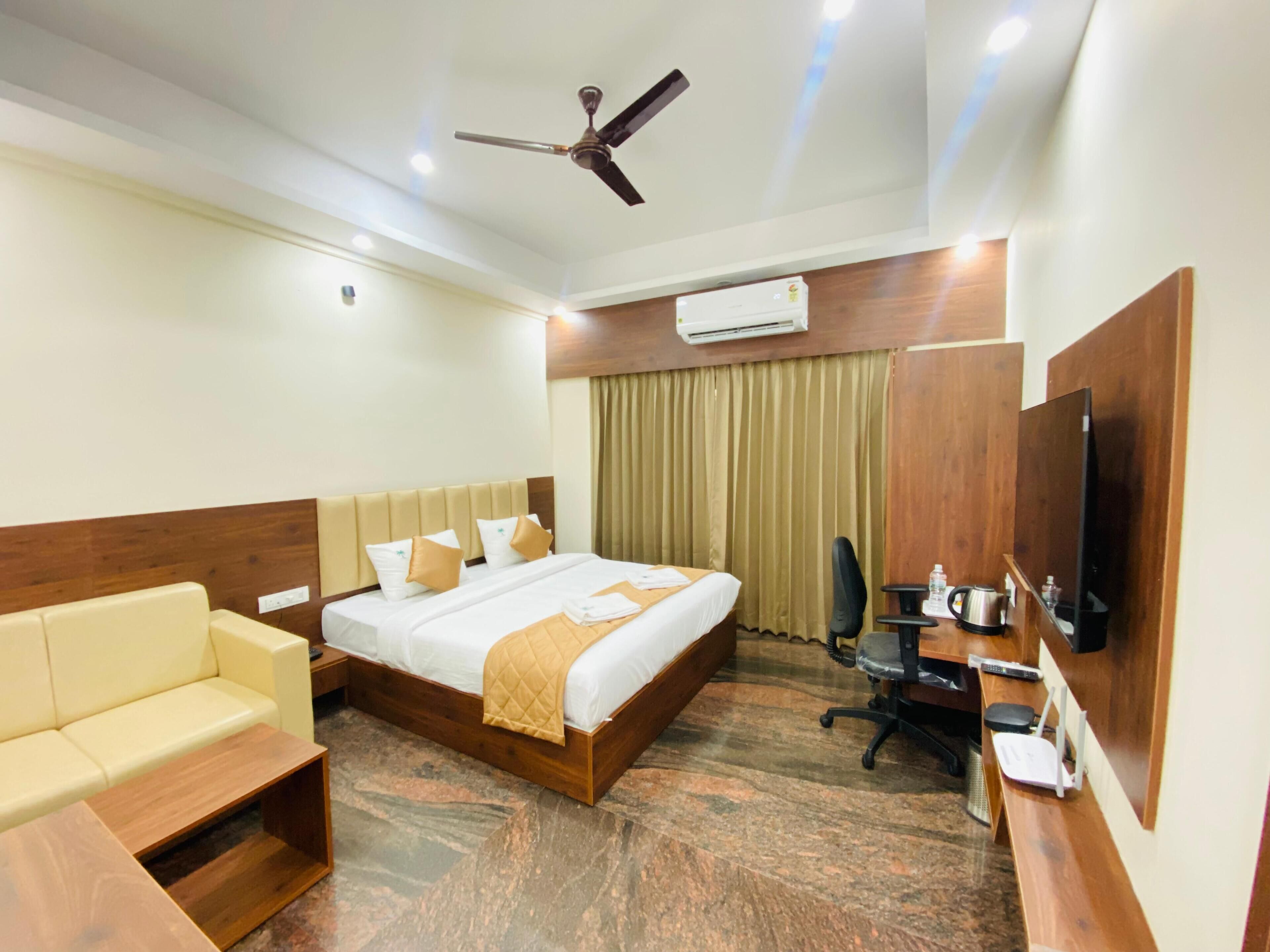 A hotel room with a bed, a sofa, a desk with a computer, a television, and an air conditioning unit.