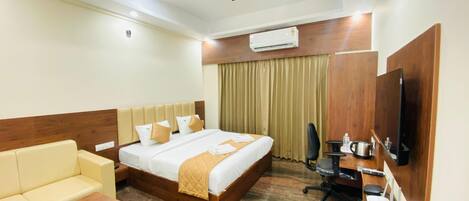 A hotel room with a bed, a sofa, a desk with a computer, a television, and an air conditioning unit.