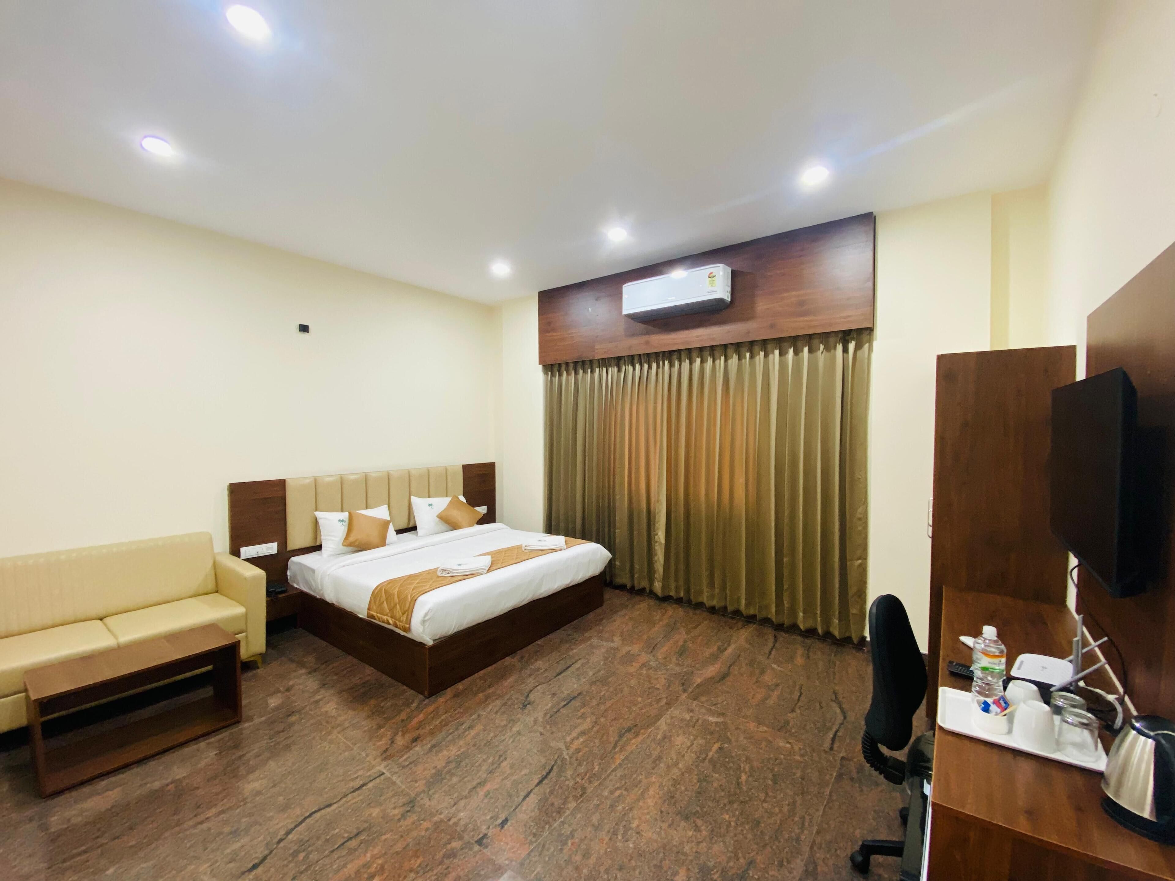 A hotel room with a bed, a sofa, a desk with a chair, an air conditioning unit, and a television.