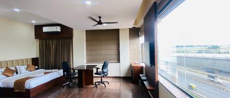 A hotel room with a bed, desk, chairs, a ceiling fan, an air conditioning unit, and a large window with a view of a highway.
