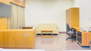 A hotel room with a wooden desk, a beige sofa, a small wooden table, a TV mounted on the wall, and a wardrobe. - Hotel Sahara Tree (Bengaluru)
