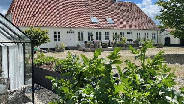A white building with a red-tiled roof, a glass greenhouse, and a garden area with green plants. - Apartment in rural idyll (Vamdrup)
